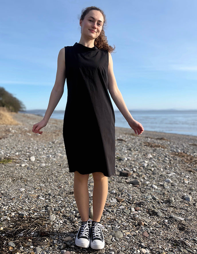 Person wearing a black dress standing on a pebbly beach with ocean and clear sky in the background
