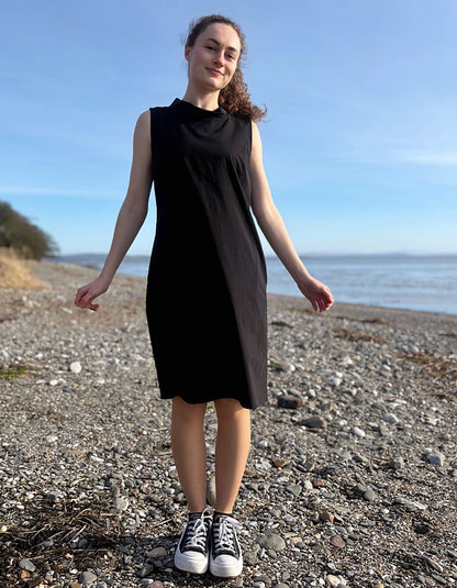 Person wearing a black dress standing on a pebbly beach with ocean and clear sky in the background