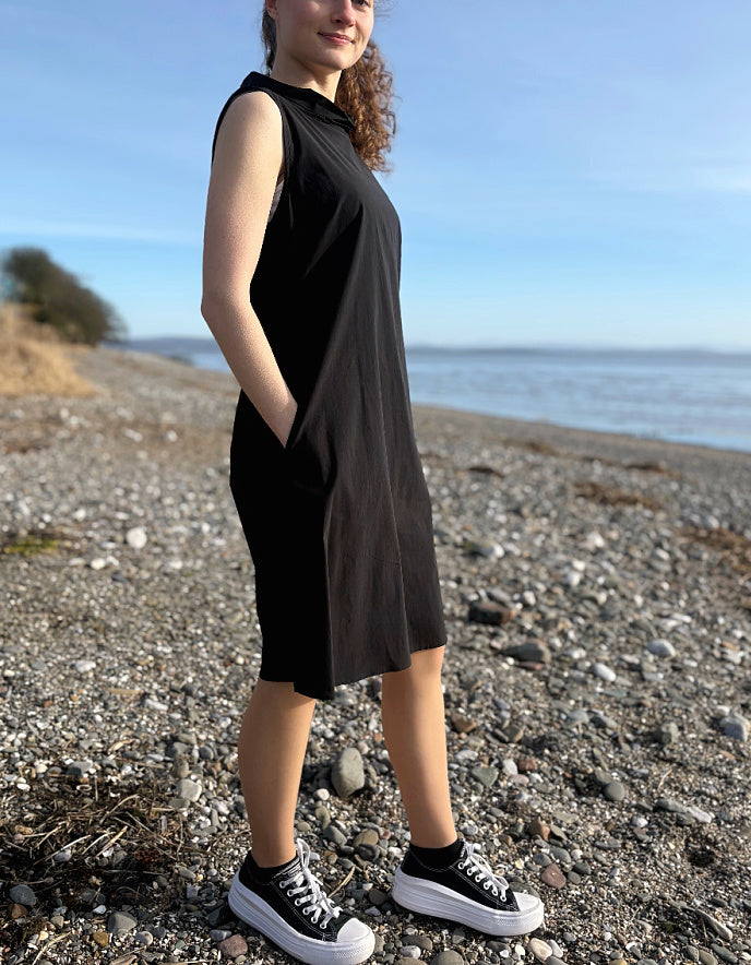 Person wearing a black sleeveless dress and black sneakers on a pebbly beach.