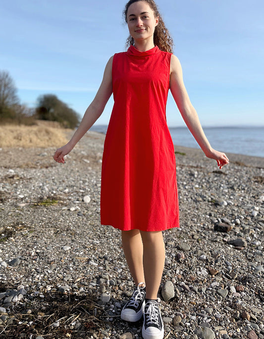 Person wearing a red dress standing on a pebbly beach with a clear sky.