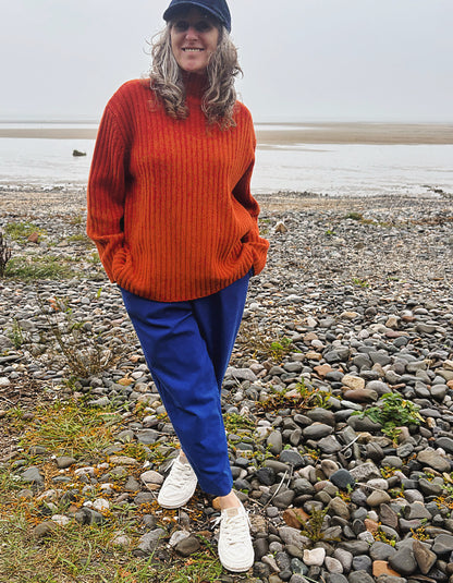 A woman standing on a rocky beach wearing a bright orange ribbed knit turtleneck sweater, blue pants, white sneakers, and a dark cap.