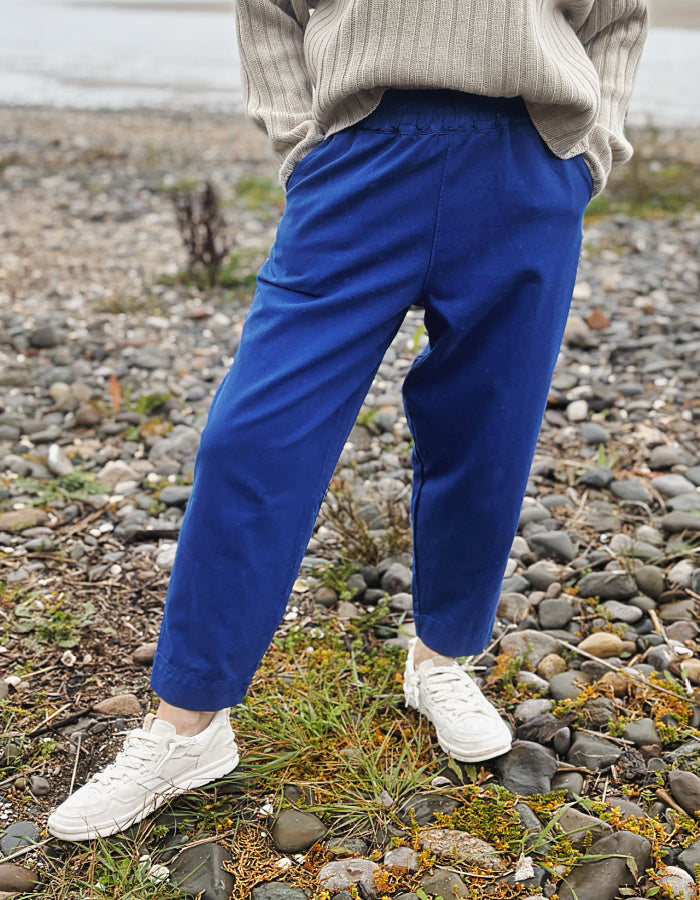 A woman standing on a rocky beach wearing bright royal blue cotton twill pants, a beige ribbed sweater, and white sneakers.