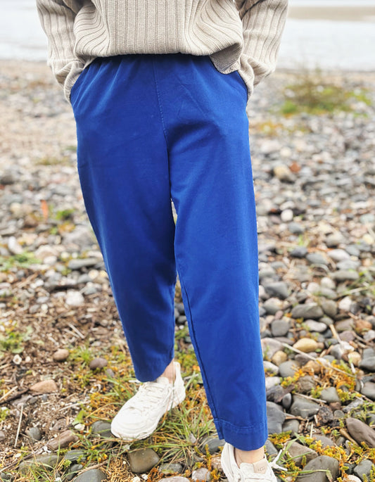 A woman standing on a rocky beach wearing bright royal blue cotton twill pants, a beige ribbed sweater, and white sneakers.
