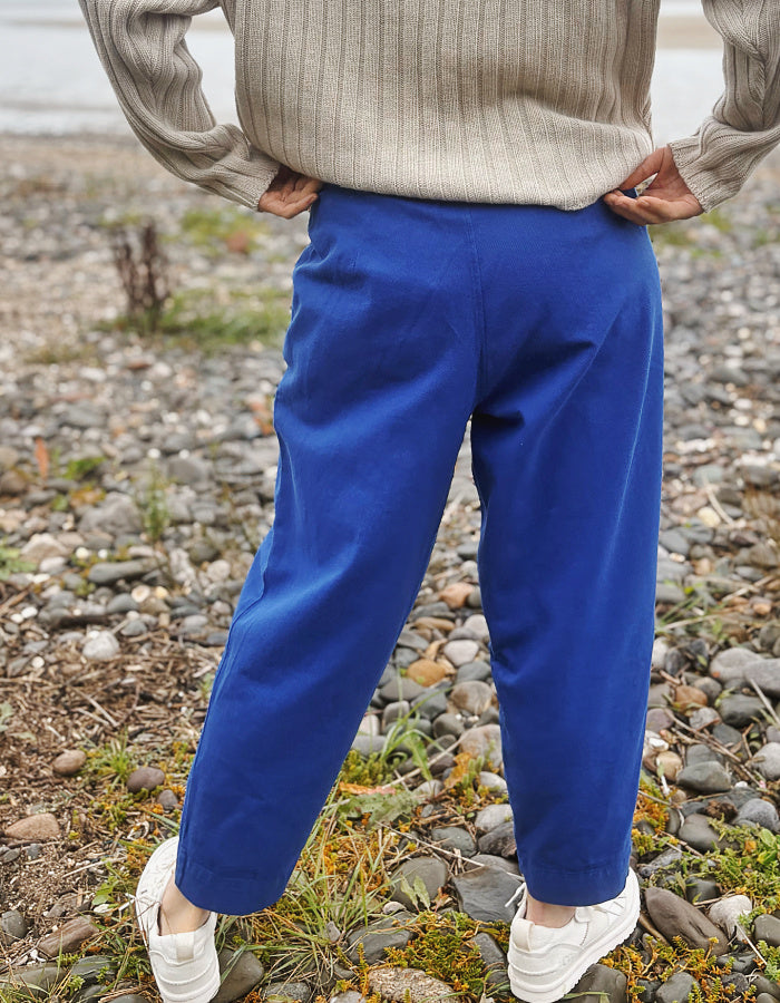 A woman standing on a rocky beach wearing bright royal blue cotton twill pants, a beige ribbed sweater, and white sneakers.