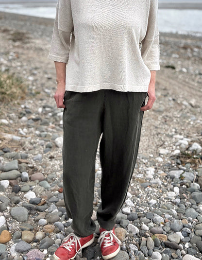Woman standing outdoors in a natural setting wearing a cream organic cotton sweater and dark green tercel pants.