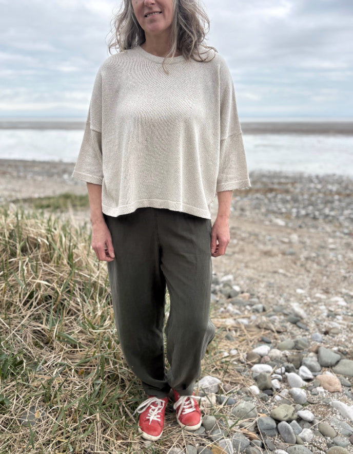 Woman standing outdoors in a natural setting wearing a cream organic cotton sweater and dark green tercel pants.