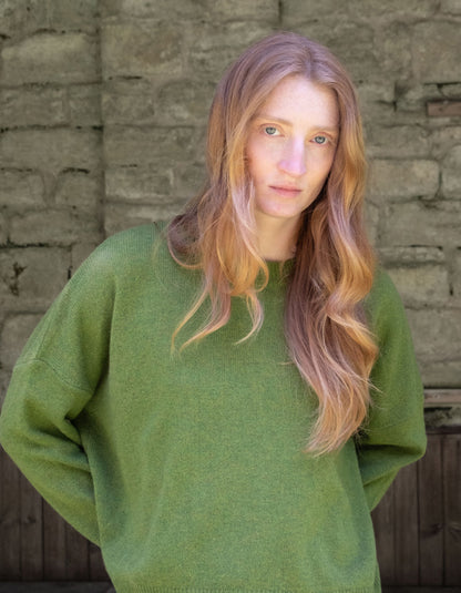 A woman with long, wavy strawberry blonde hair wearing an oversized sage green merino knit sweater, standing in front of a rustic stone wall.