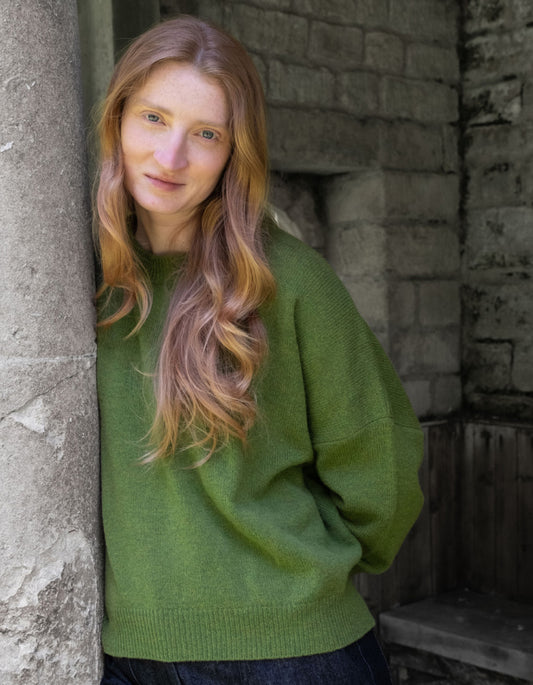 A woman with long wavy light brown hair wearing an oversized olive green knit sweater, leaning against a rustic stone pillar in an outdoor setting.