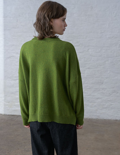 A woman with shoulder-length brown hair is shown from the back wearing an oversized olive green knit sweater and dark trousers, standing against a white brick wall.