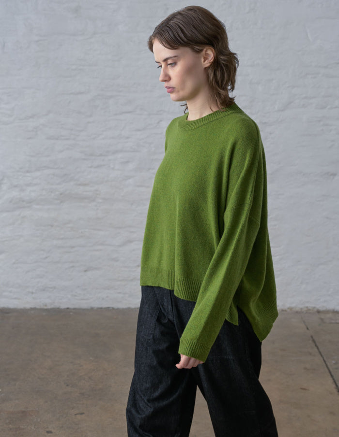 A woman wearing a bright olive green knit sweater and dark wide-leg trousers, standing in profile against a light gray textured wall and concrete floor.