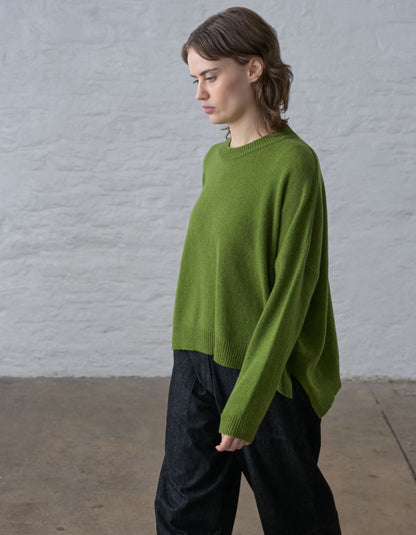 A woman wearing a bright olive green knit sweater and dark wide-leg trousers, standing in profile against a light gray textured wall and concrete floor.