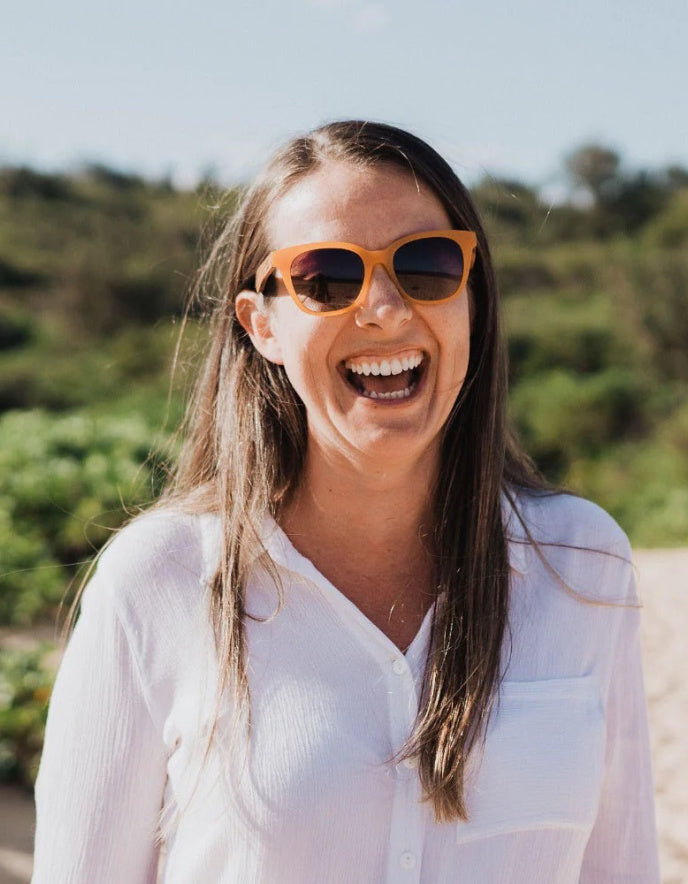 A woman with long brown hair wearing orange-framed sunglasses and a white button-up shirt, smiling outdoors with a blurred green and blue background.