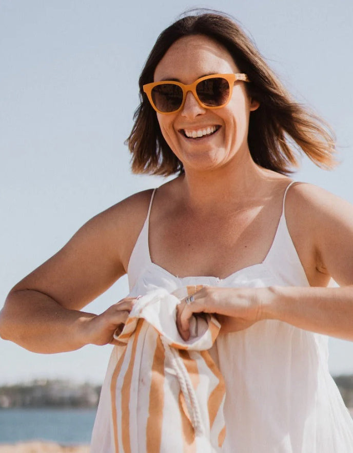 A woman outdoors wearing orange-framed sunglasses and a white spaghetti-strap dress, holding a white and orange striped towel. The background shows a bright sky and distant water.