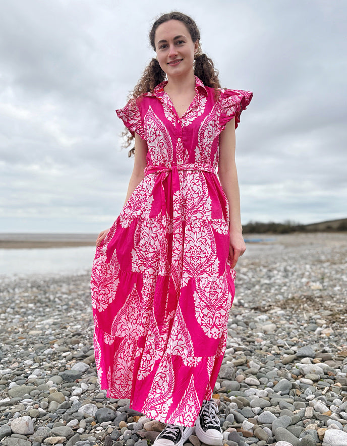Woman in a pink and white patterned dress standing on a pebbly beach.