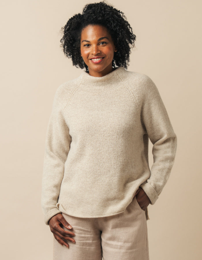 A woman with curly black hair wearing a light beige knit mock-neck sweater and matching beige trousers, standing against a solid beige background.