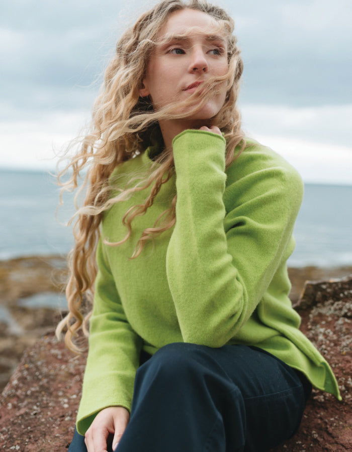 A woman with long wavy blonde hair sits on a rocky shore wearing a bright lime green knit sweater and dark navy pants, with the ocean and cloudy sky in the background.