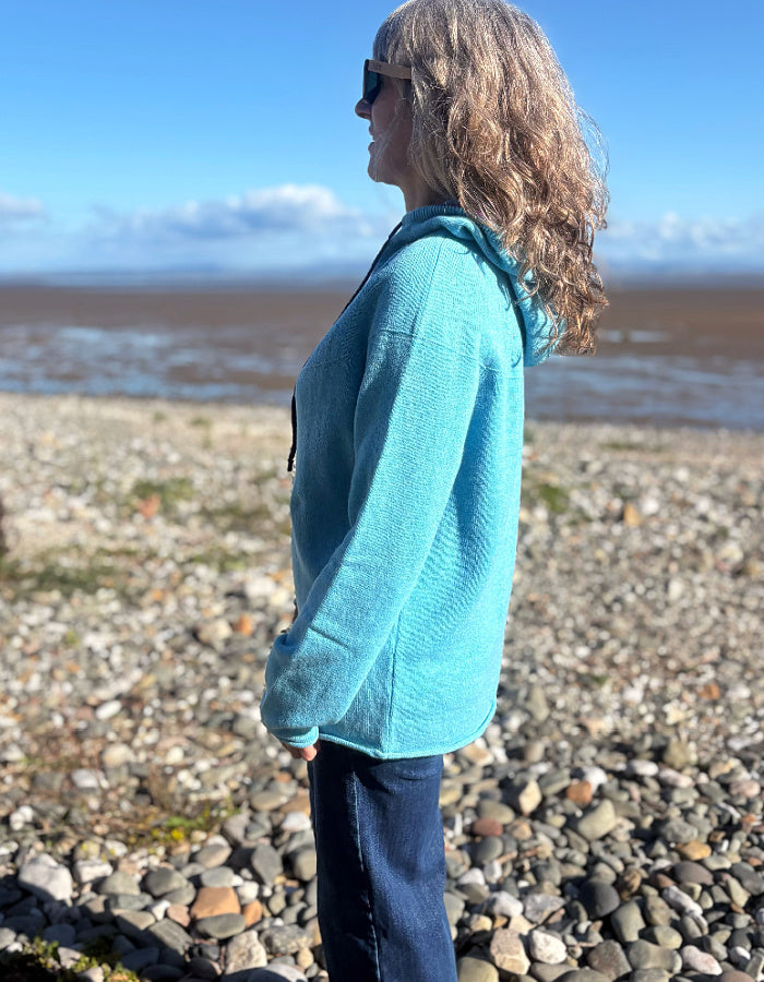 A woman with curly blonde hair stands in profile on a rocky beach, wearing a bright turquoise fleece hoodie and dark blue jeans under a blue sky.