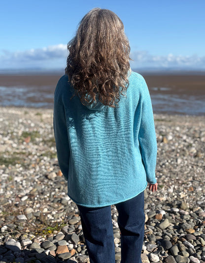 A person with long curly brown hair stands on a rocky beach facing the water, wearing a textured light blue knit sweater and dark jeans.