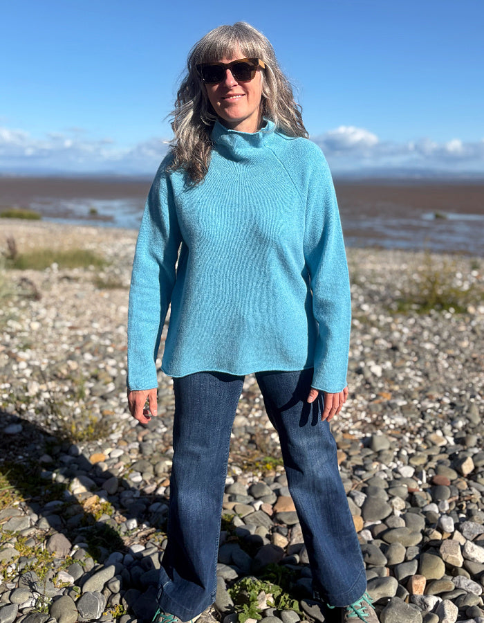 A woman wearing a bright turquoise ribbed turtleneck sweater and dark blue jeans stands on a rocky beach with water and a blue sky in the background.