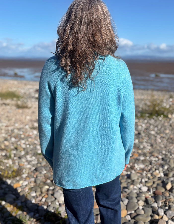 A person viewed from behind wearing a heathered turquoise blue knit sweater and dark jeans, standing on a pebble beach with water and sky in the background.