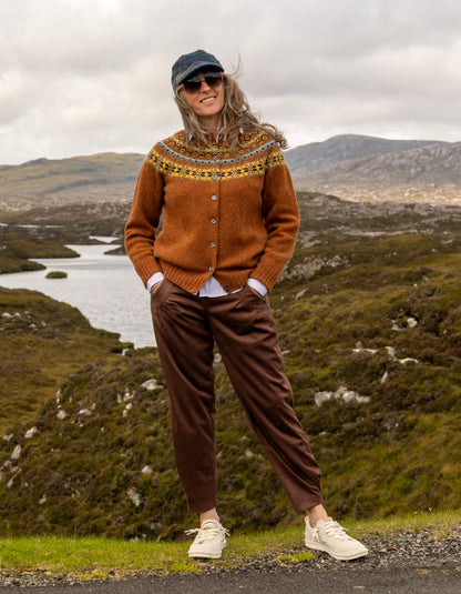 A woman stands outdoors in a rugged mountain landscape wearing a rust-brown Fair Isle cardigan, brown trousers, white sneakers, and a dark blue cap.