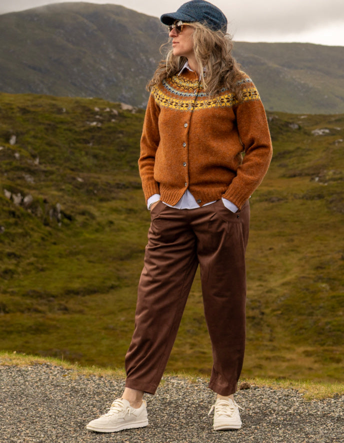 A woman stands outdoors in a grassy mountain landscape wearing a rust-orange knit cardigan with a multicolor yoke pattern, brown trousers, white sneakers, and a blue cap.