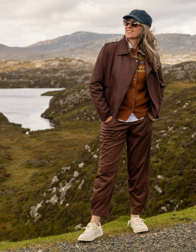 A woman stands outdoors in a rugged mountain landscape wearing a brown tailored jacket and matching trousers, layered over a rust-orange vest, a white shirt, and white sneakers. She is also wearing a dark blue cap and sunglasses.