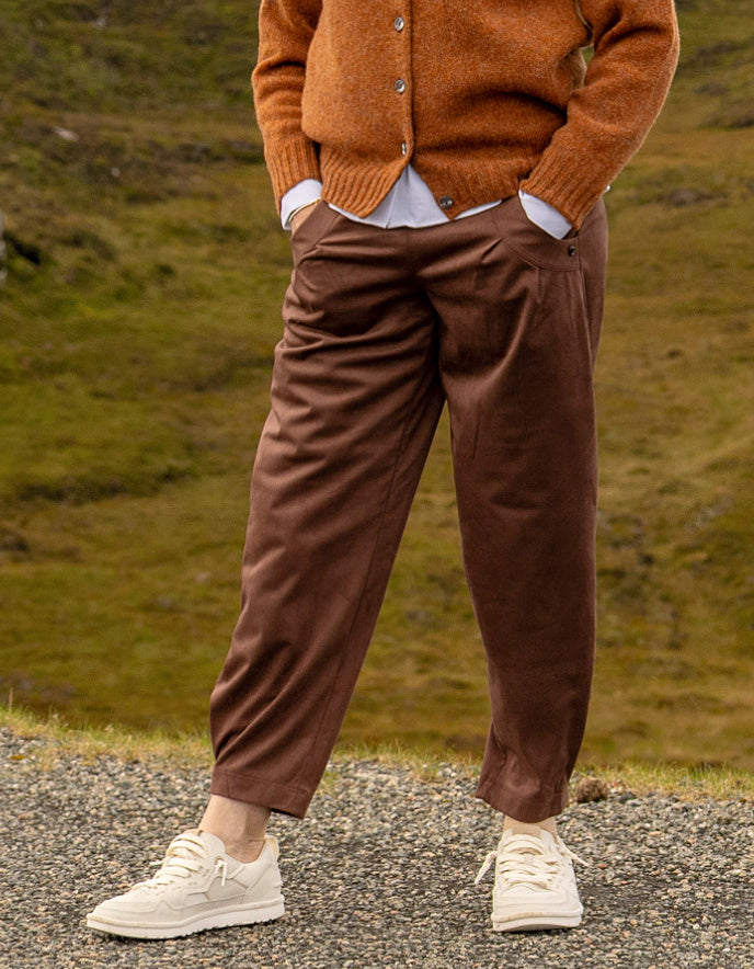 A person standing outdoors on a gravel path wearing brown tapered pants, a rust-orange cardigan, a white shirt, and off-white sneakers, with a blurred grassy landscape in the background.