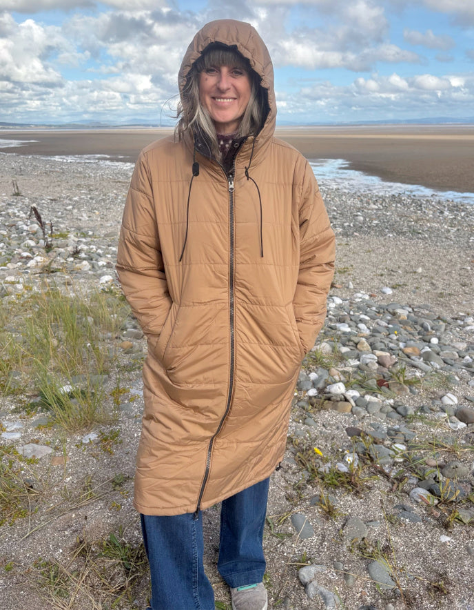 Person wearing a long beige coat on a beach with a cloudy sky.