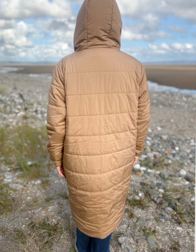 Person wearing a long beige quilted coat standing on a rocky beach with a cloudy sky.