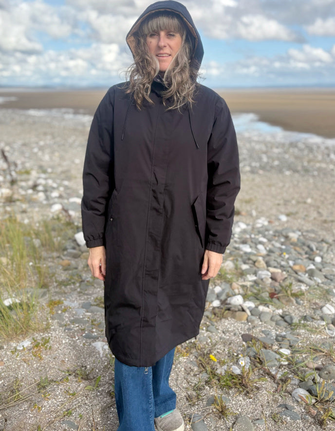Person wearing a black coat standing on a rocky beach with a cloudy sky.