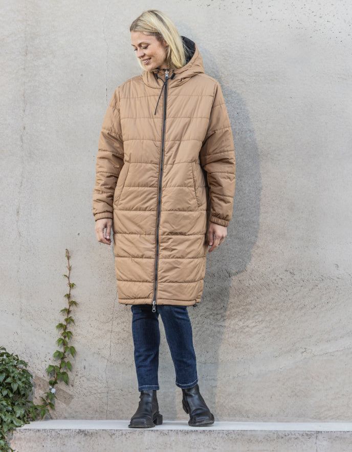 Woman wearing a long quilted beige coat against a plain wall.