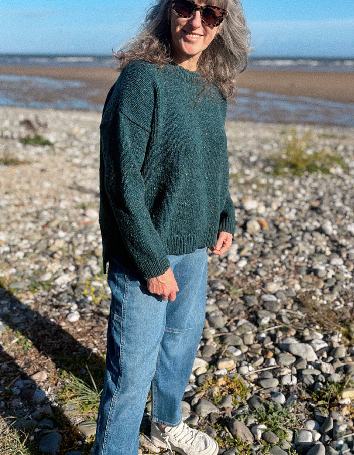 A woman wearing a dark green donegal tweed knit crewneck sweater and blue jeans stands on a rocky beach with the sea and blue sky in the background.