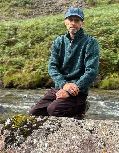 A man wearing a dark teal quarter-zip fleece sweater, dark brown pants, and a blue cap sits on a large mossy rock next to a flowing stream with green ferns in the background.