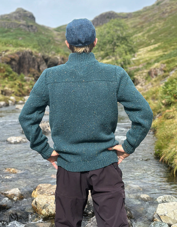 A man standing outdoors by a rocky stream, viewed from behind, wearing a textured teal-green knit sweater and a blue flat cap, with green hills in the background.