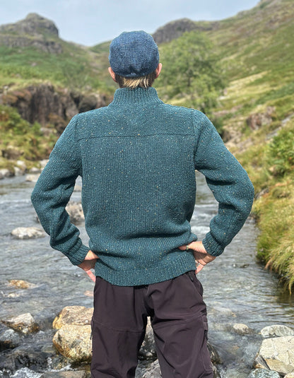 A man standing outdoors by a rocky stream, viewed from behind, wearing a textured teal-green knit sweater and a blue flat cap, with green hills in the background.