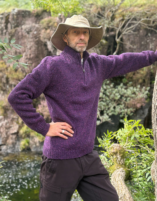 A man wearing a textured purple quarter-zip sweater, dark cargo pants, and a beige wide-brim hat, posing outdoors in front of a rocky, green landscape.