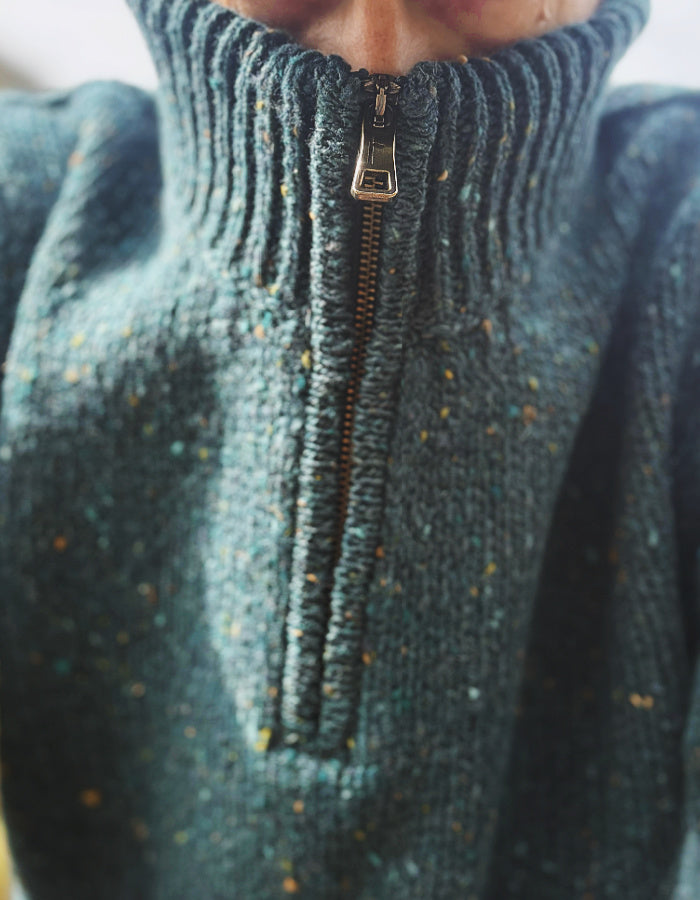 Close-up of a ribbed knit half-zip sweater in a speckled teal green yarn with small multicolored flecks, featuring a silver metal zipper pull.