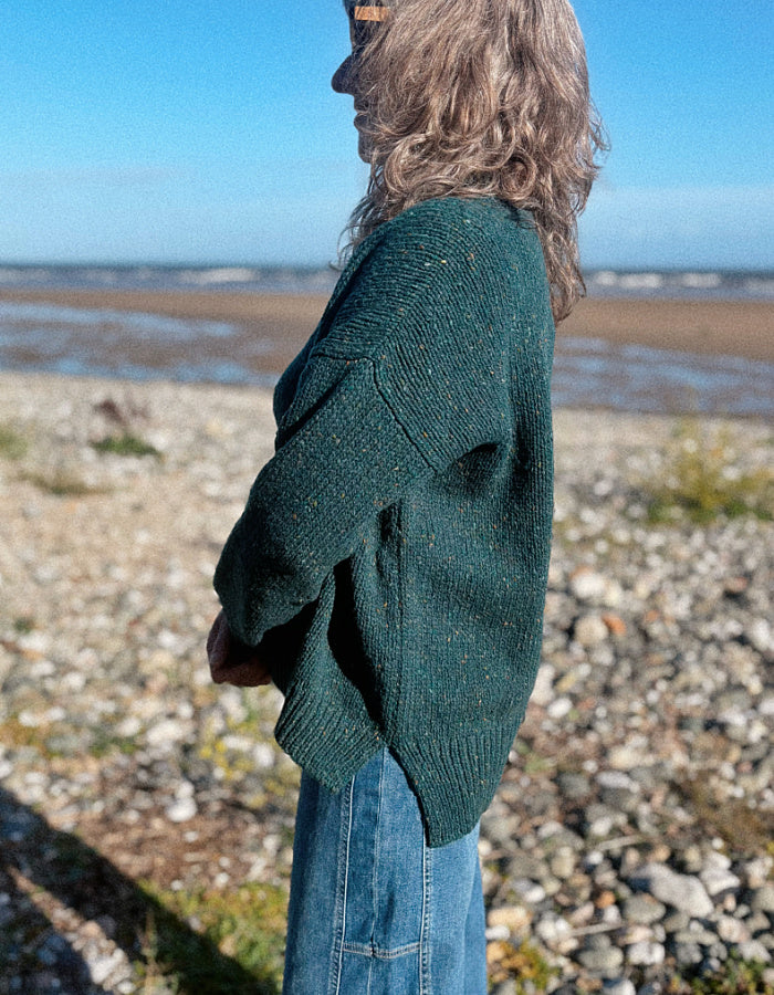 A woman wearing a dark green donegal tweed knit crewneck sweater and blue jeans stands on a rocky beach with the sea and blue sky in the background.