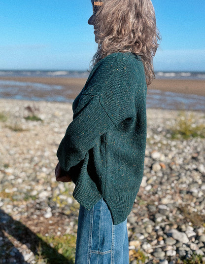 A woman wearing a dark green donegal tweed knit crewneck sweater and blue jeans stands on a rocky beach with the sea and blue sky in the background.