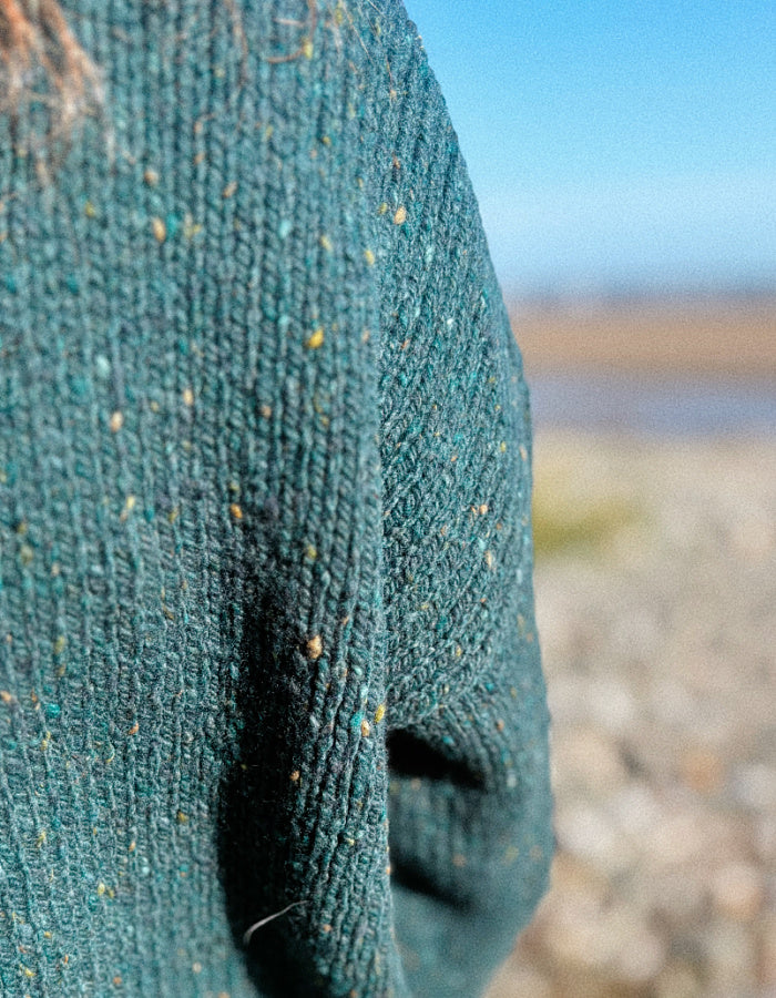 Close-up of a textured knit sweater in a deep teal green with flecks of yellow, blue, and orange, shown outdoors with a blurred beach and sky background.