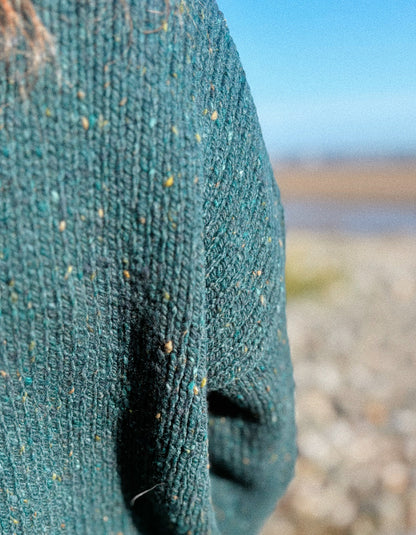 Close-up of a textured knit sweater in a deep teal green with flecks of yellow, blue, and orange, shown outdoors with a blurred beach and sky background.