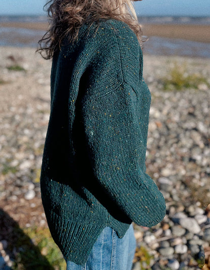 A person standing outdoors on a pebble beach wearing an oversized dark green knit sweater with subtle multicolor flecks and blue jeans.