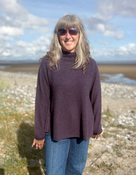 Woman wearing a purple yak wool swing shaped sweater and sunglasses standing on a beach with a blue sky and clouds in the background.