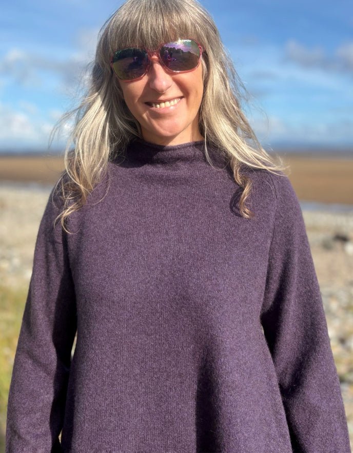 Woman wearing a purple yak wool swing shaped sweater and sunglasses standing on a beach with a blue sky and clouds in the background.