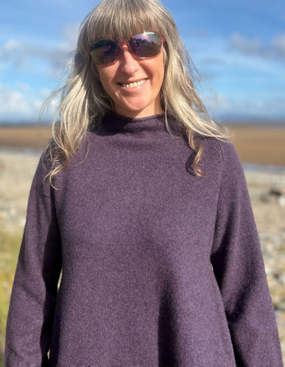 Woman wearing a purple yak wool swing shaped sweater and sunglasses standing on a beach with a blue sky and clouds in the background.