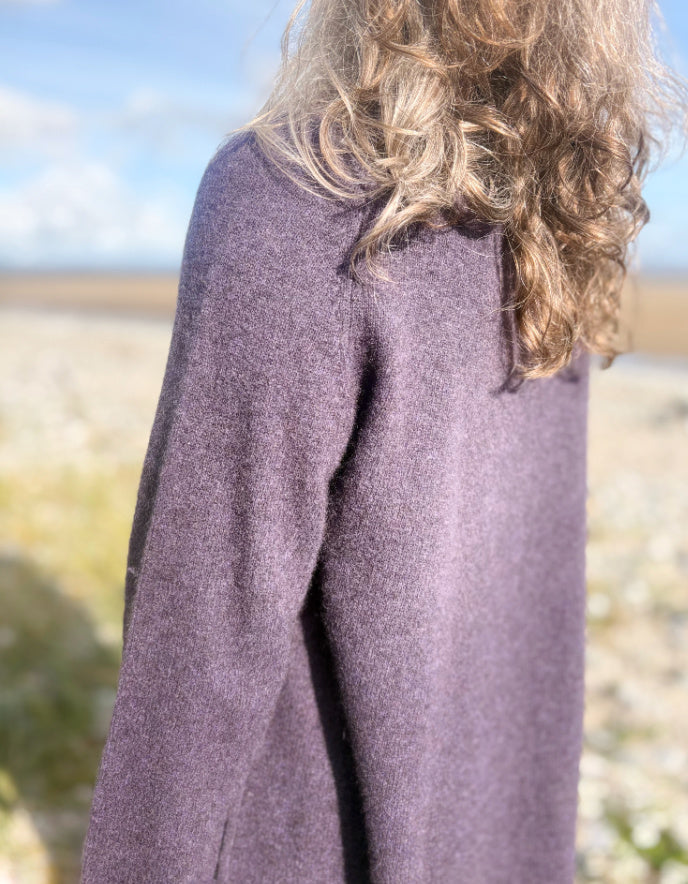 Woman wearing a purple yak wool swing shaped sweater and sunglasses standing on a beach with a blue sky and clouds in the background.