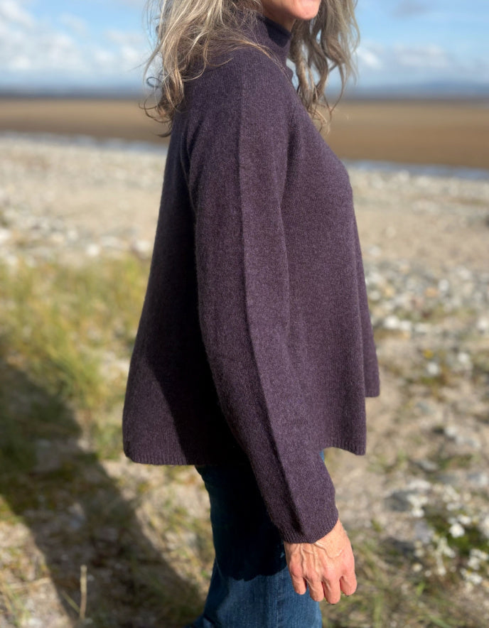 Woman wearing a purple yak wool swing shaped sweater and sunglasses standing on a beach with a blue sky and clouds in the background.