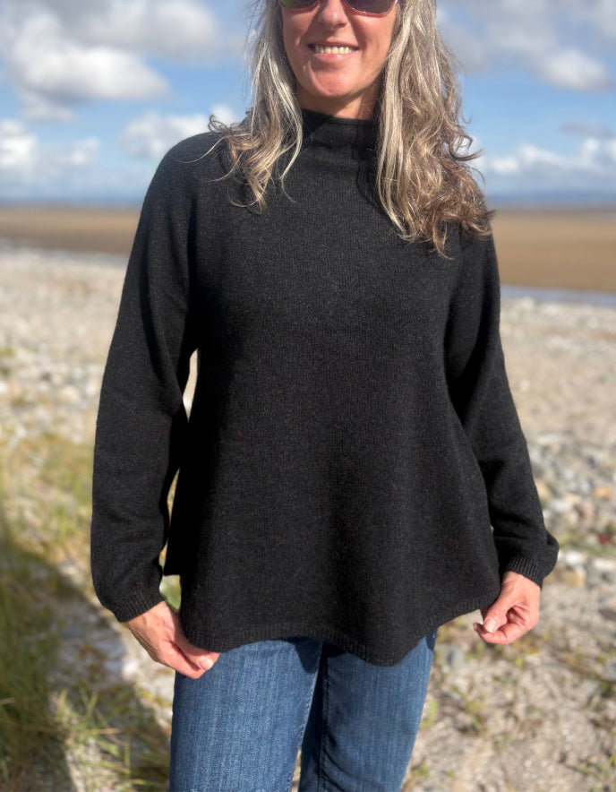 Person wearing a black yak wool sweater and blue jeans standing on a beach with a scenic background.
