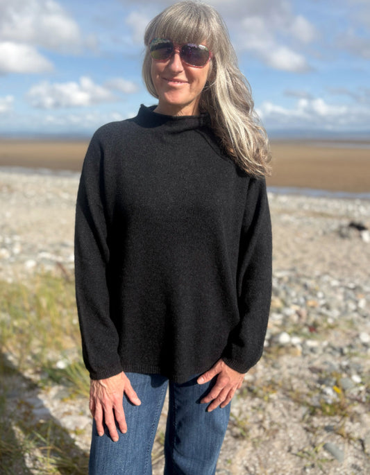 Person wearing a black yak wool sweater and blue jeans standing on a beach with a scenic background.